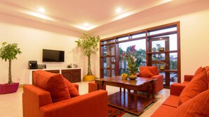 A modern living room with orange sofas, a wooden coffee table, a flat-screen TV, and large windows with a view of greenery. - Eco Villa (Siem Reap)
