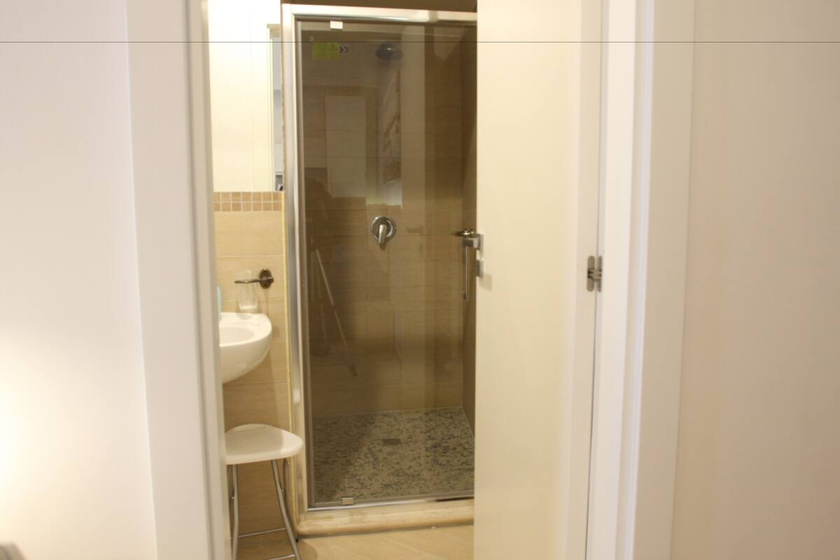 Un baño con ducha de vidrio, un lavamanos blanco y un taburete blanco pequeño.