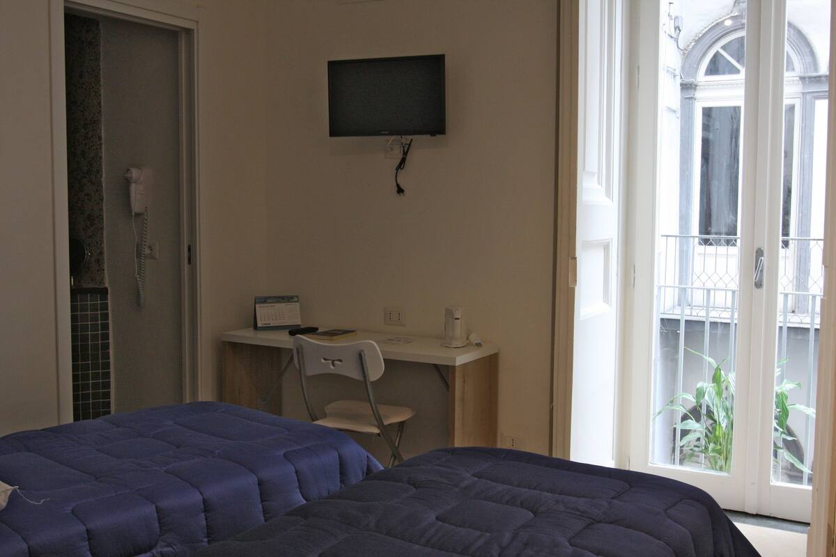 Habitación de hotel con dos camas, un escritorio con silla, un televisor en la pared y una ventana con vista al exterior.