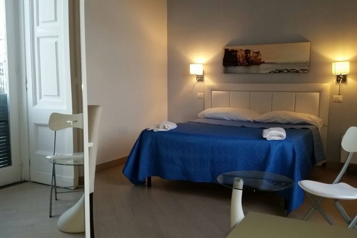 Habitación de hotel con cama, dos sillas, una mesita y dos lámparas fijadas a la pared.