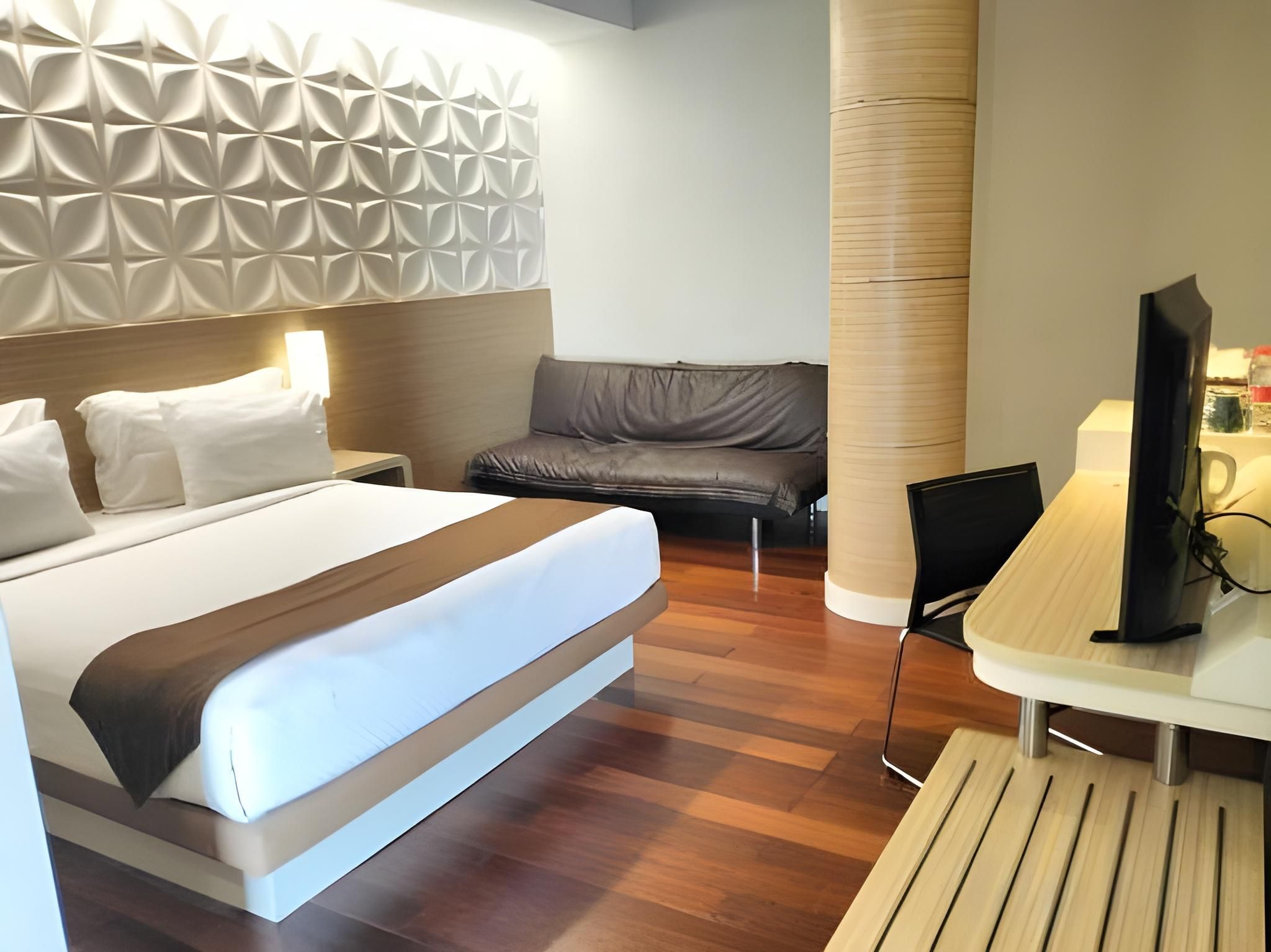 A modern hotel room with a bed, a seating area, a desk with a computer, and a television.