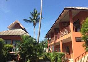 Un edificio de dos pisos con un balcón en el nivel superior, rodeado de abundante vegetación y palmeras.