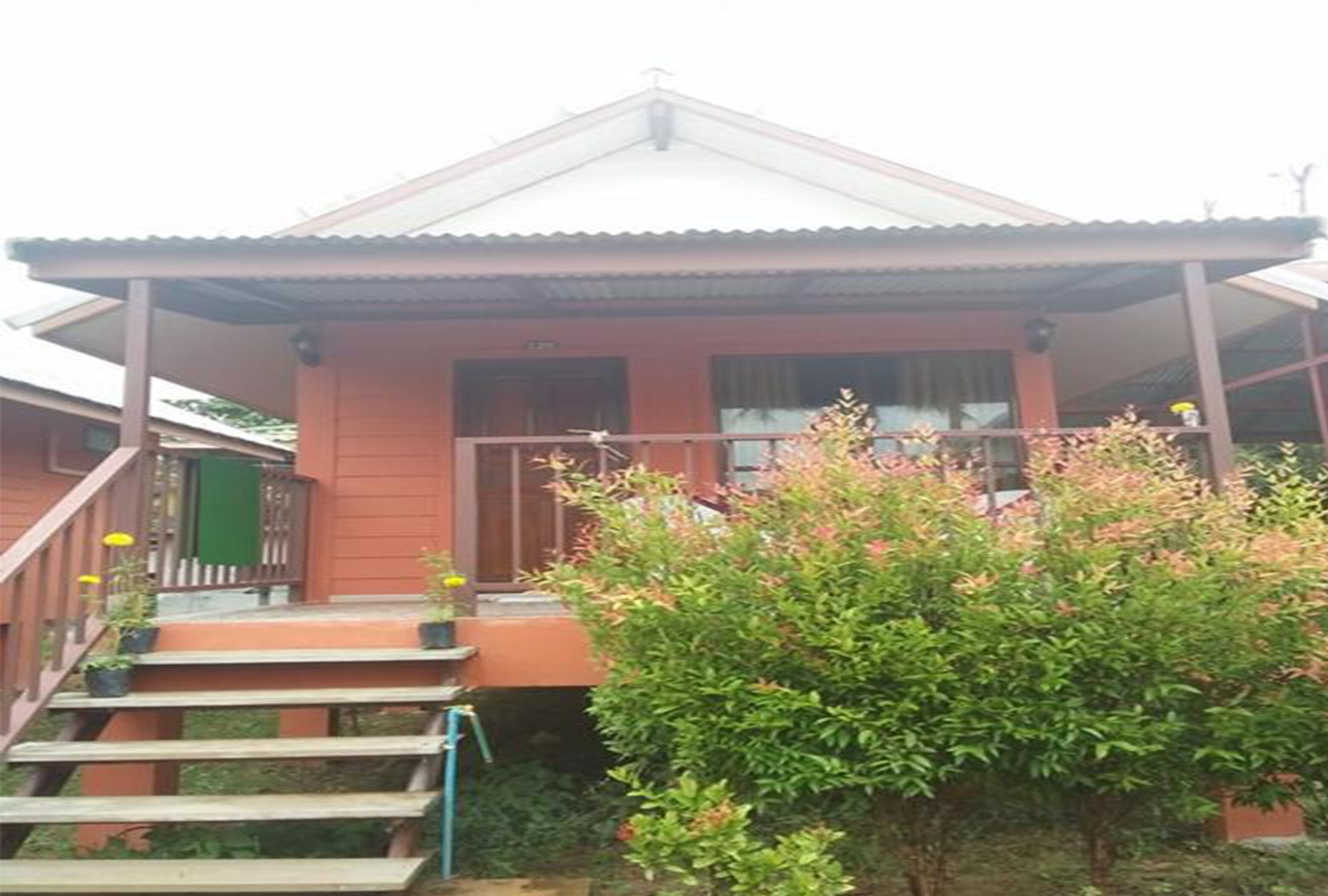 Una casa de dos pisos con fachada roja, un porche cubierto y un pequeño jardín con un arbusto y flores amarillas.