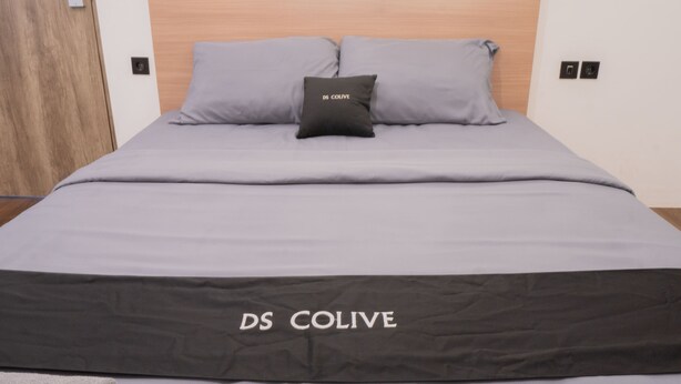 A bed with grey bedding and a black pillow with "DS COLIVE" embroidered on it. - DS Residence Sinabung (Semarang)