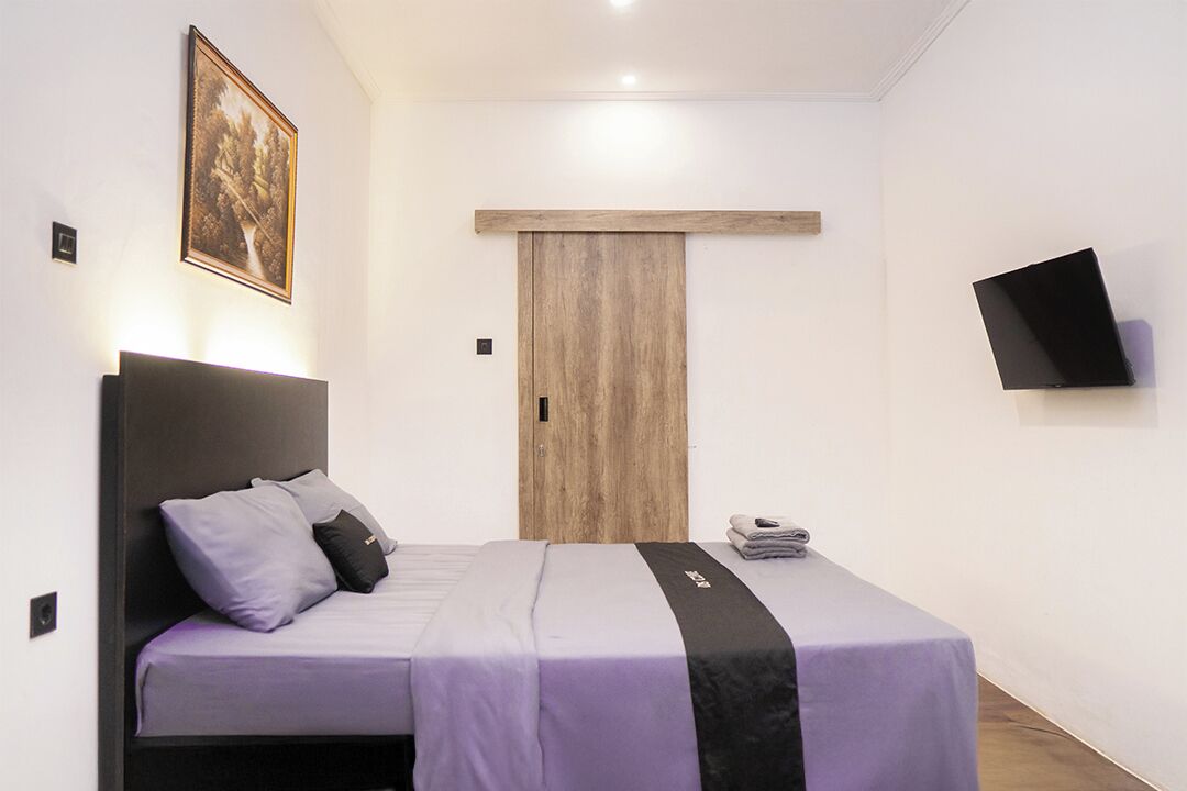 Et moderne hotelværelse med en seng, en trædør, et ophængt fjernsyn og et billede i ramme på væggen.