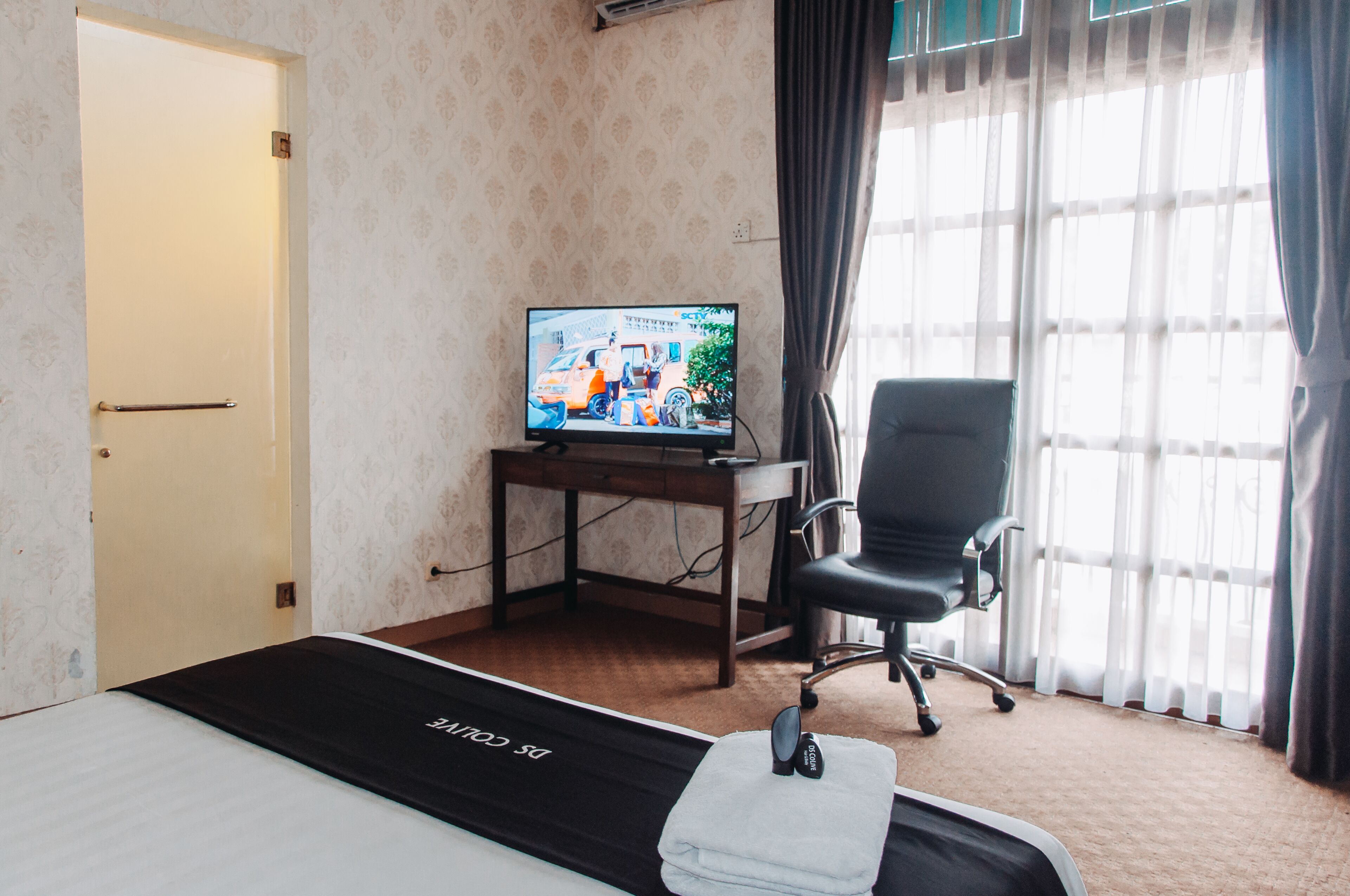A hotel room with a bed, a desk with a television, a chair, and a window with curtains.