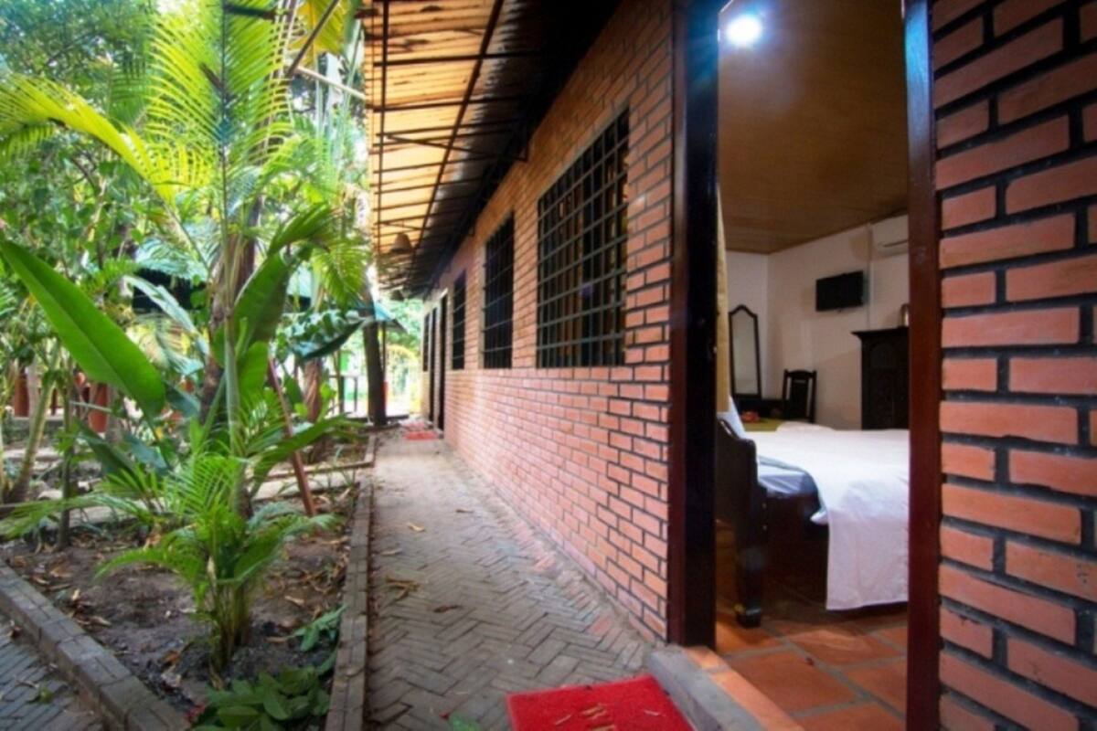 Un edificio de ladrillo con un corredor cubierto, una cama con ropa de cama blanca y un pequeño jardín con plantas.