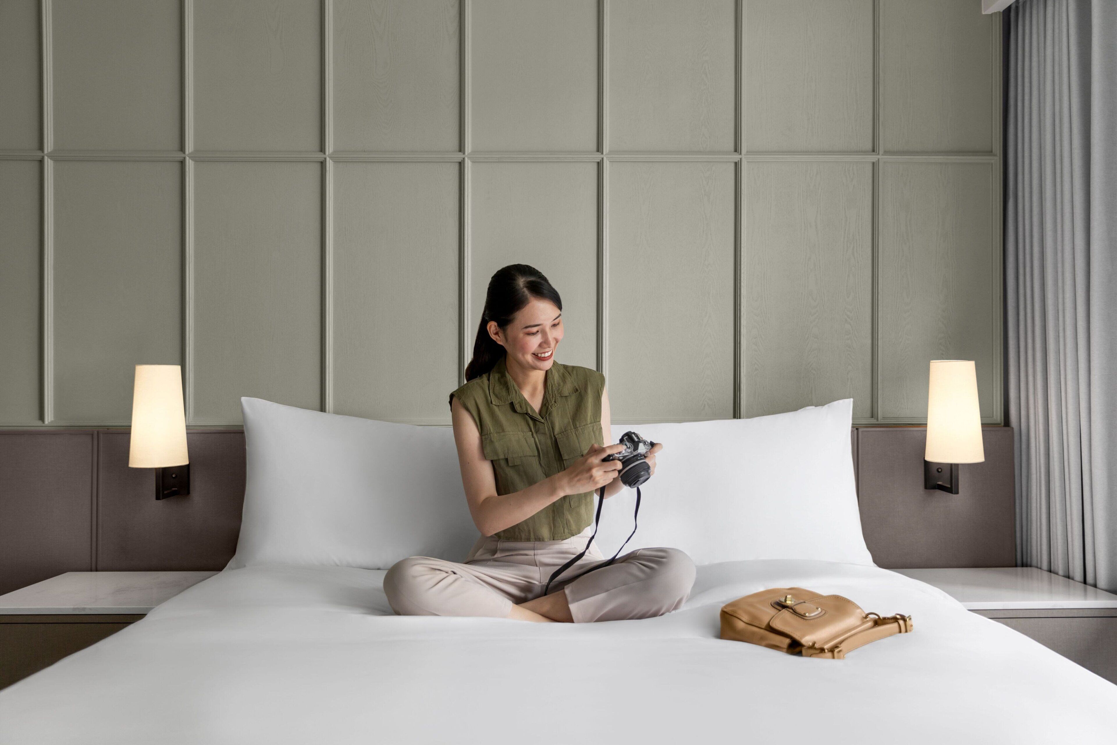 A person sitting on a bed in a hotel room with a camera, a lamp, and a handbag on the bed.