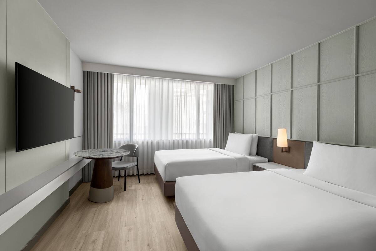 A modern hotel room with two beds, a flat-screen TV, a round dining table, and a window with curtains.
