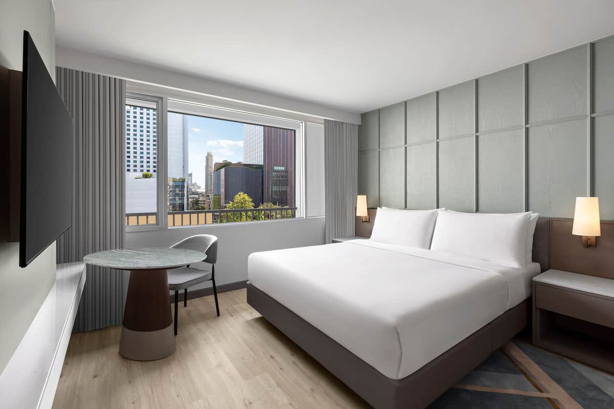 A modern hotel room with a large bed, a round table, and a chair. There is a window with a view of buildings and a cityscape.