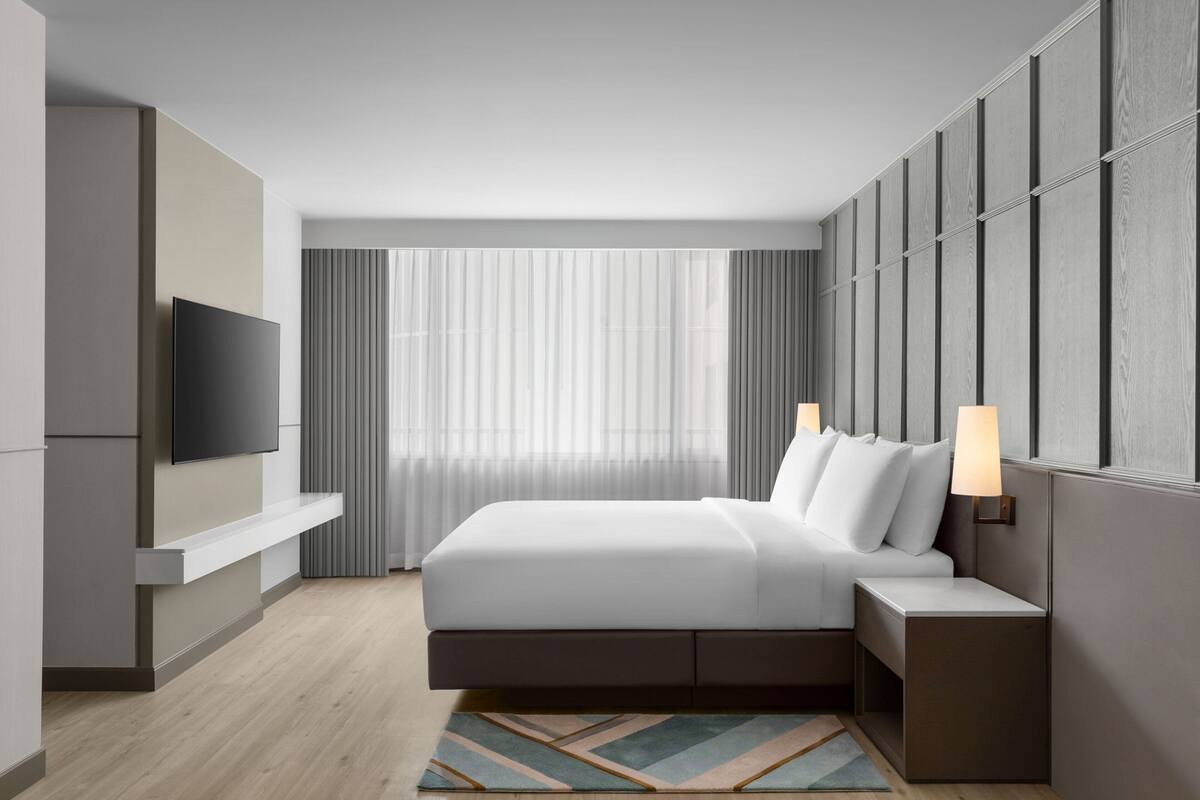 A modern hotel room with a large bed, a TV mounted on the wall, and a window with sheer curtains.