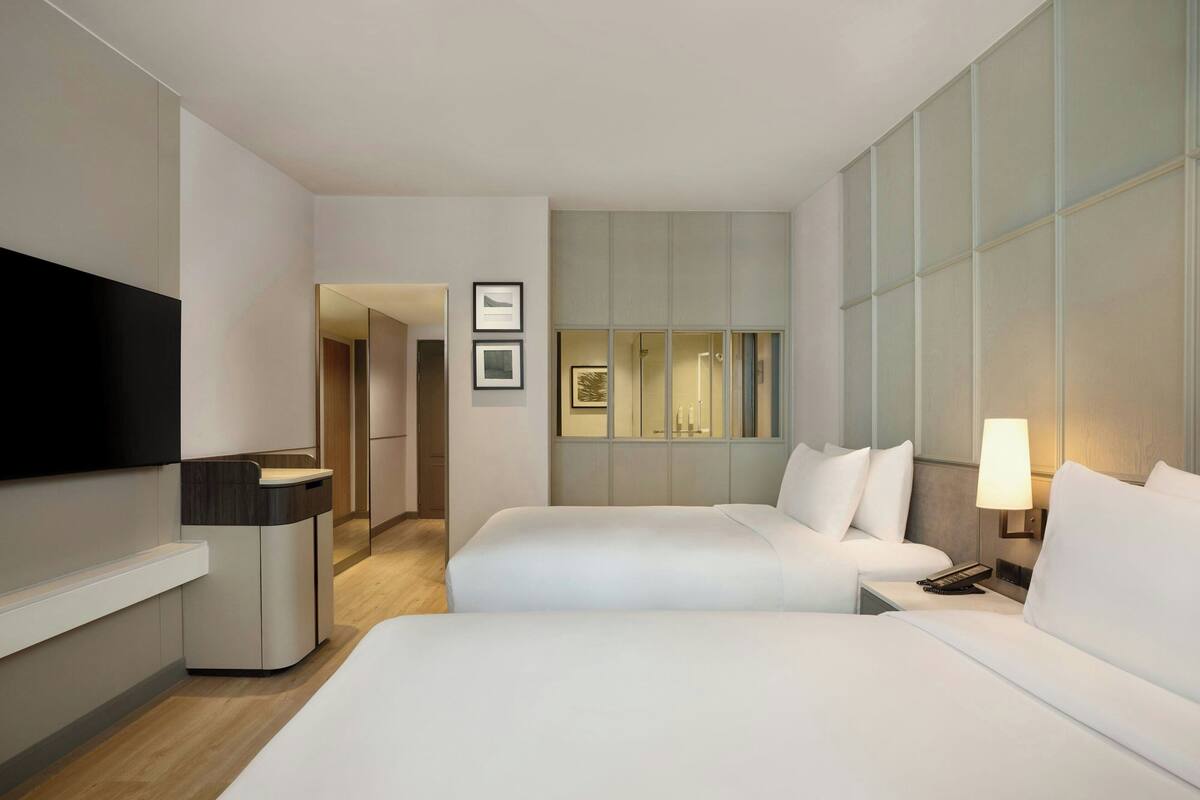 A modern hotel room with two beds, a TV, a bedside table with a lamp, and a mirror.
