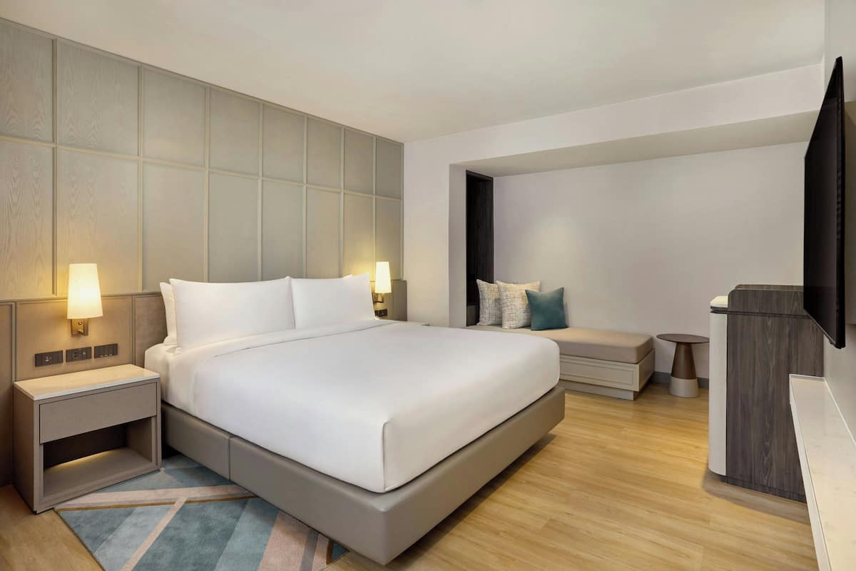A modern hotel room with a large bed, bedside tables with lamps, a small seating area, and a TV on the wall.