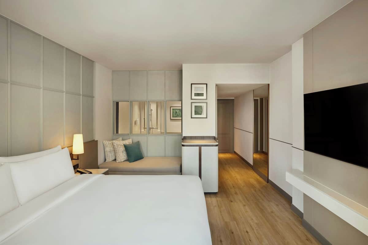 A modern hotel room with a large bed, a bedside table with a lamp, a small seating area with a bench, a kitchenette, and a TV mounted on the wall.