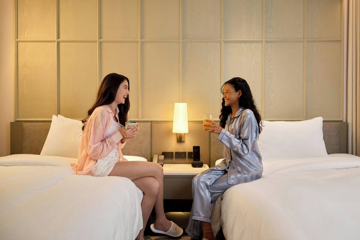 Two people in pajamas on a hotel bed with a nightstand, a lamp, and a phone.