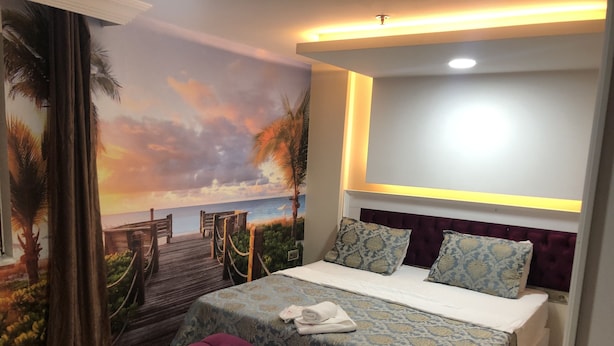 A bedroom with a bed, a bench, a mural of a sunset beach scene, and a headboard with built-in lighting. - Sanli Hotel (Trabzon)