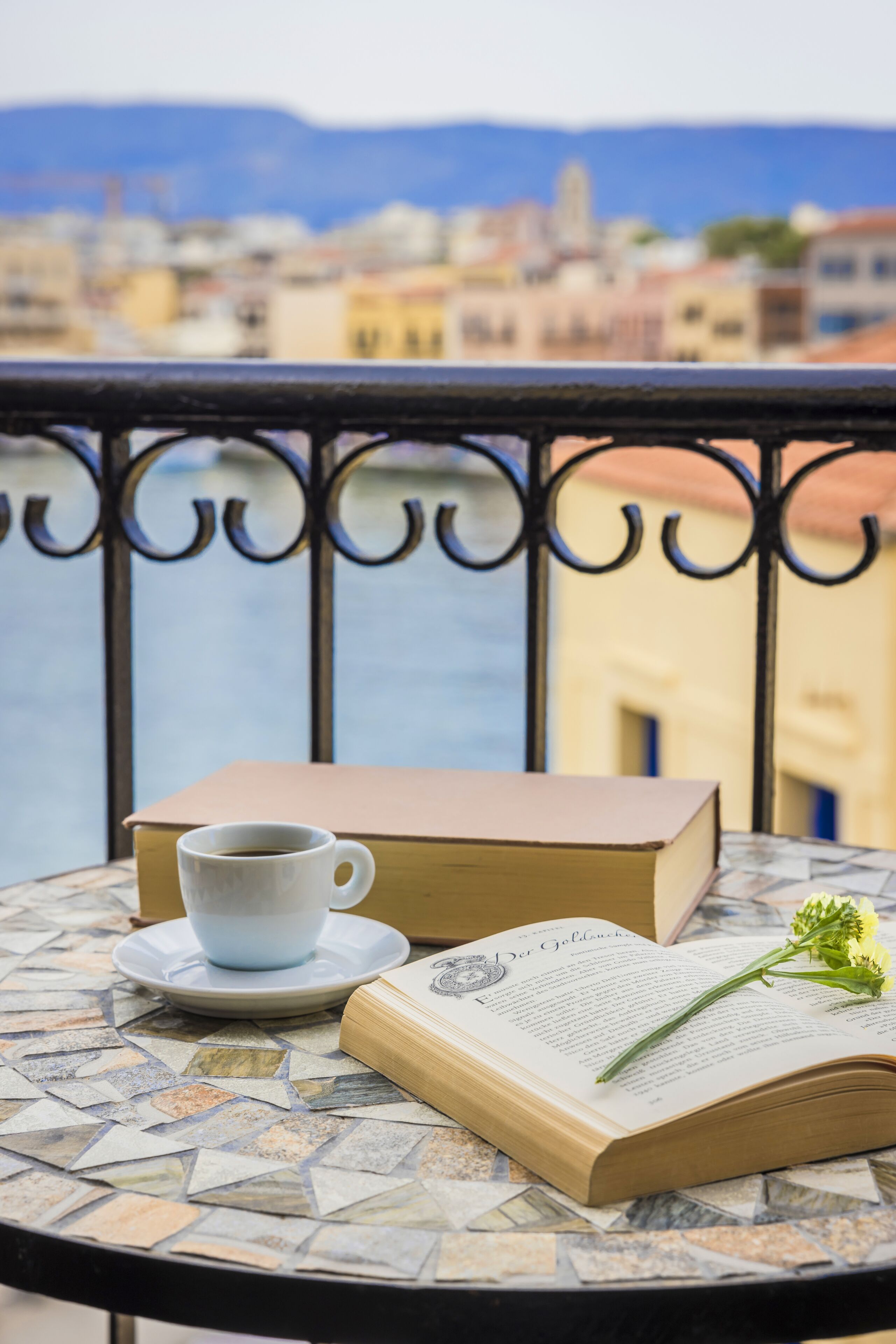 A table with a book, a cup, and a flower on a tiled balcony overlooking a cityscape.
