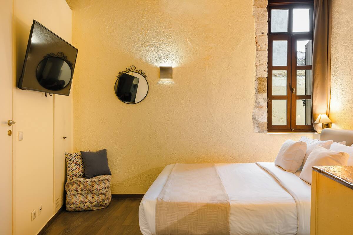 Un dormitorio con una cama, una mesita de noche pequeña, una mesita auxiliar, un televisor montado en la pared, un espejo y una ventana con cortinas.