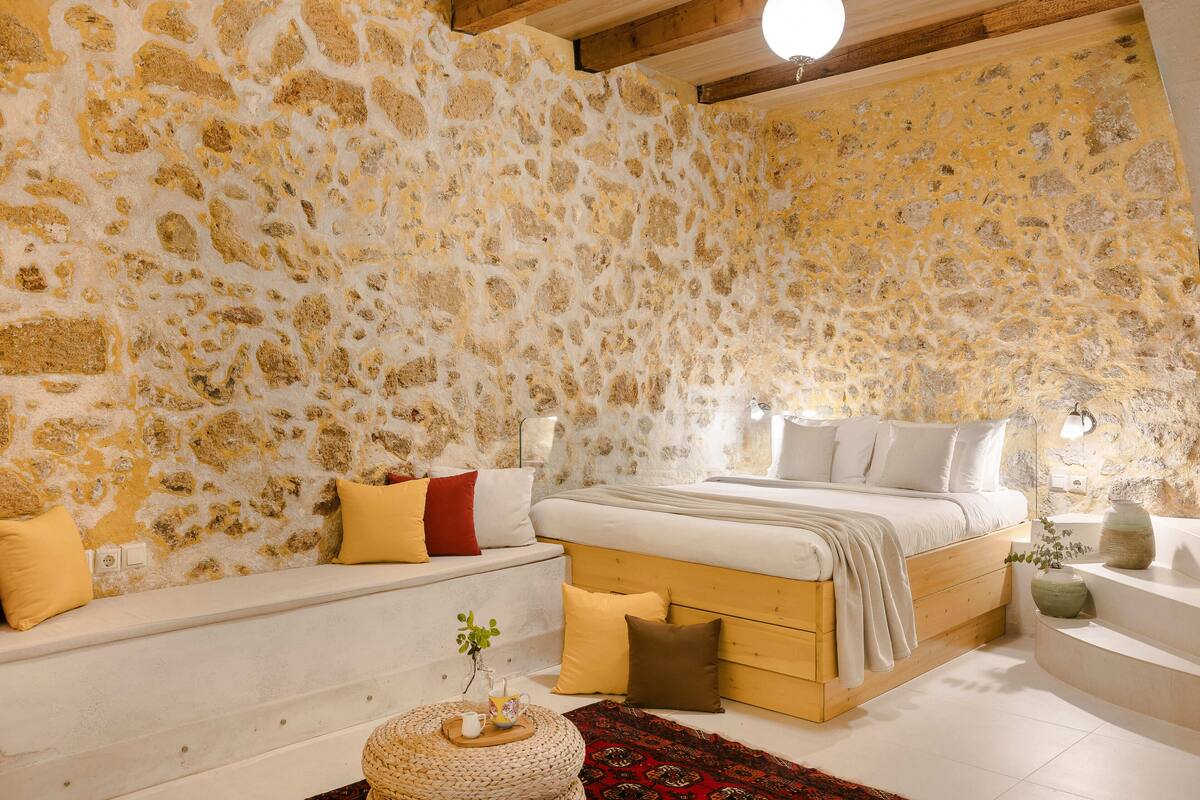 Un dormitorio con pared de piedra, cama con ropa de cama blanca, un banco de madera y una pequeña mesa con una planta.