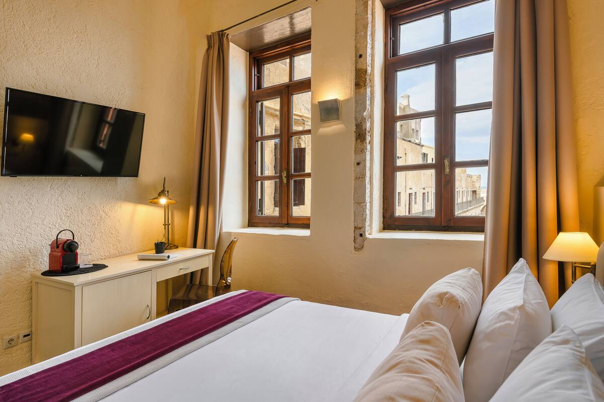 Habitación de hotel con cama, escritorio, televisor y ventana con vista a edificios.