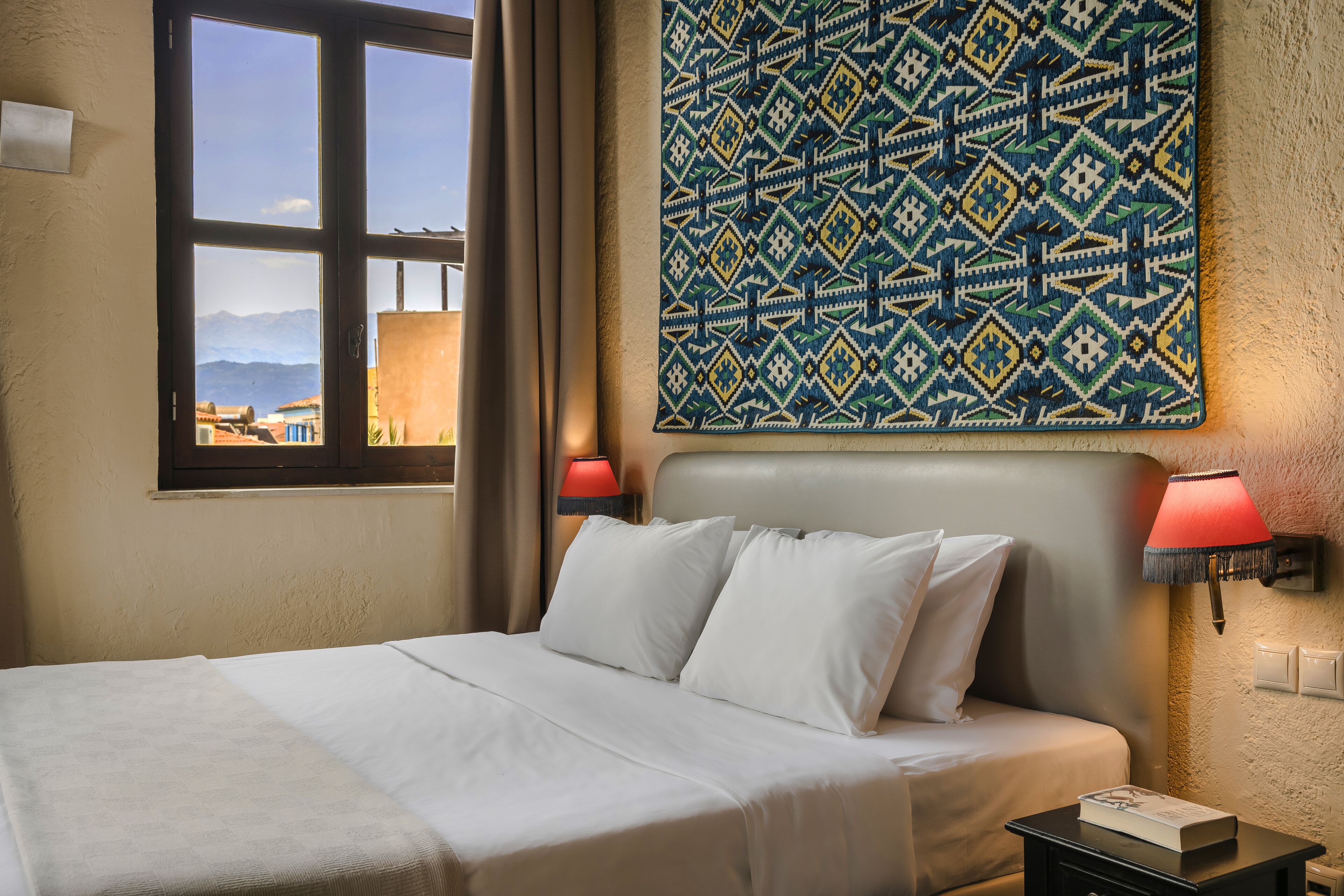 Una camera d'albergo con un letto, due abat-jour, un pannello decorativo a parete e una finestra che si affaccia su edifici e montagne.
