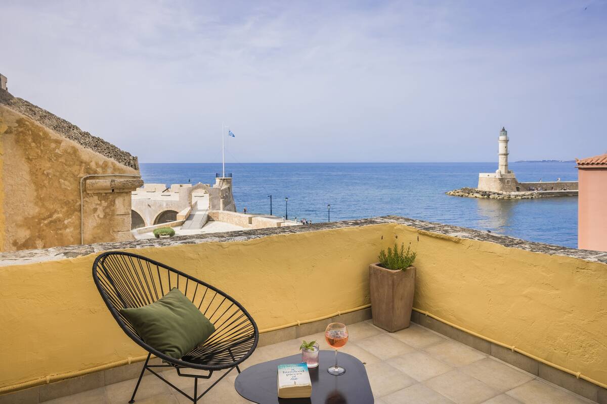 Un balcón con vista al mar, una silla, una mesa con un libro y una copa de vino, y una planta en maceta.