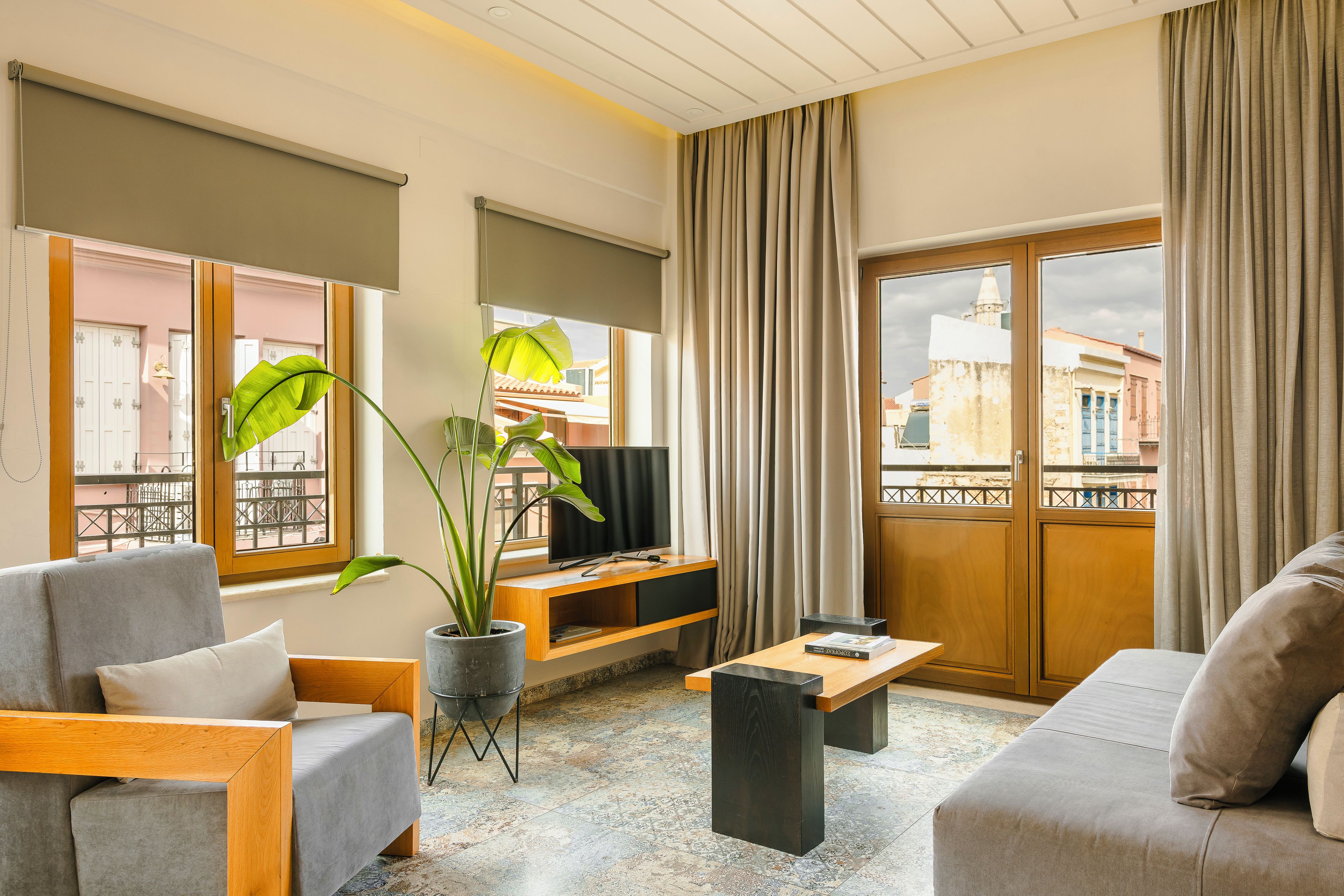 Una sala de estar moderna con un sofá, una mesa de centro y un televisor sobre un mueble de madera. Hay un ventanal con cortinas, un balcón y una planta.