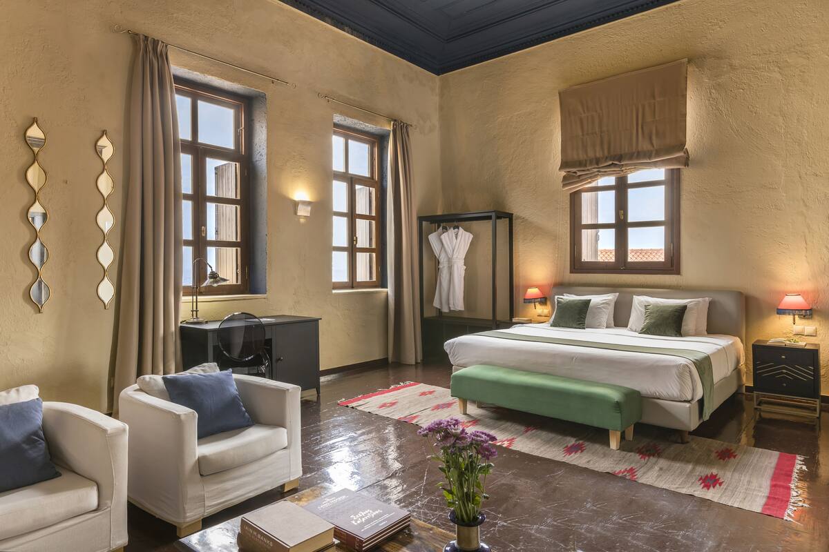 Un dormitorio bien iluminado con una cama, mesitas de noche, un escritorio, una silla y una ventana con cortinas.