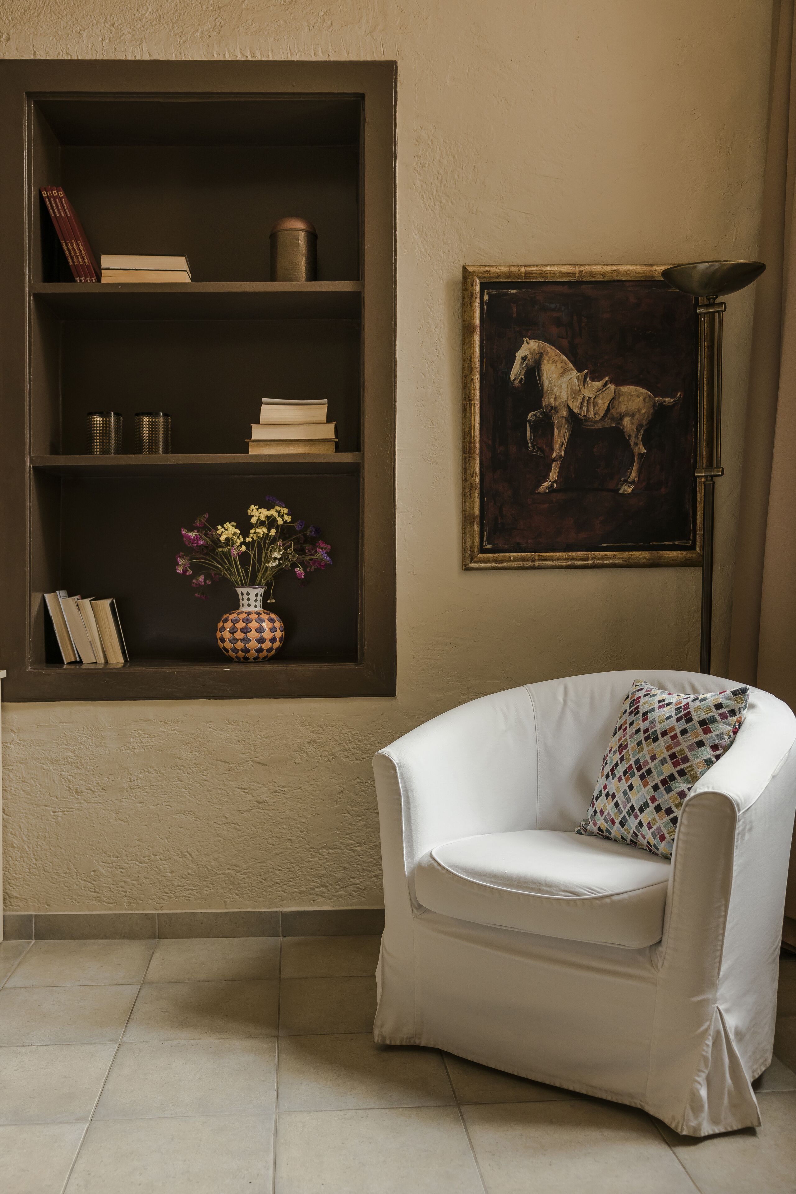 A room with a white armchair, a bookshelf with books and decorative items, and a framed painting of a horse on the wall.