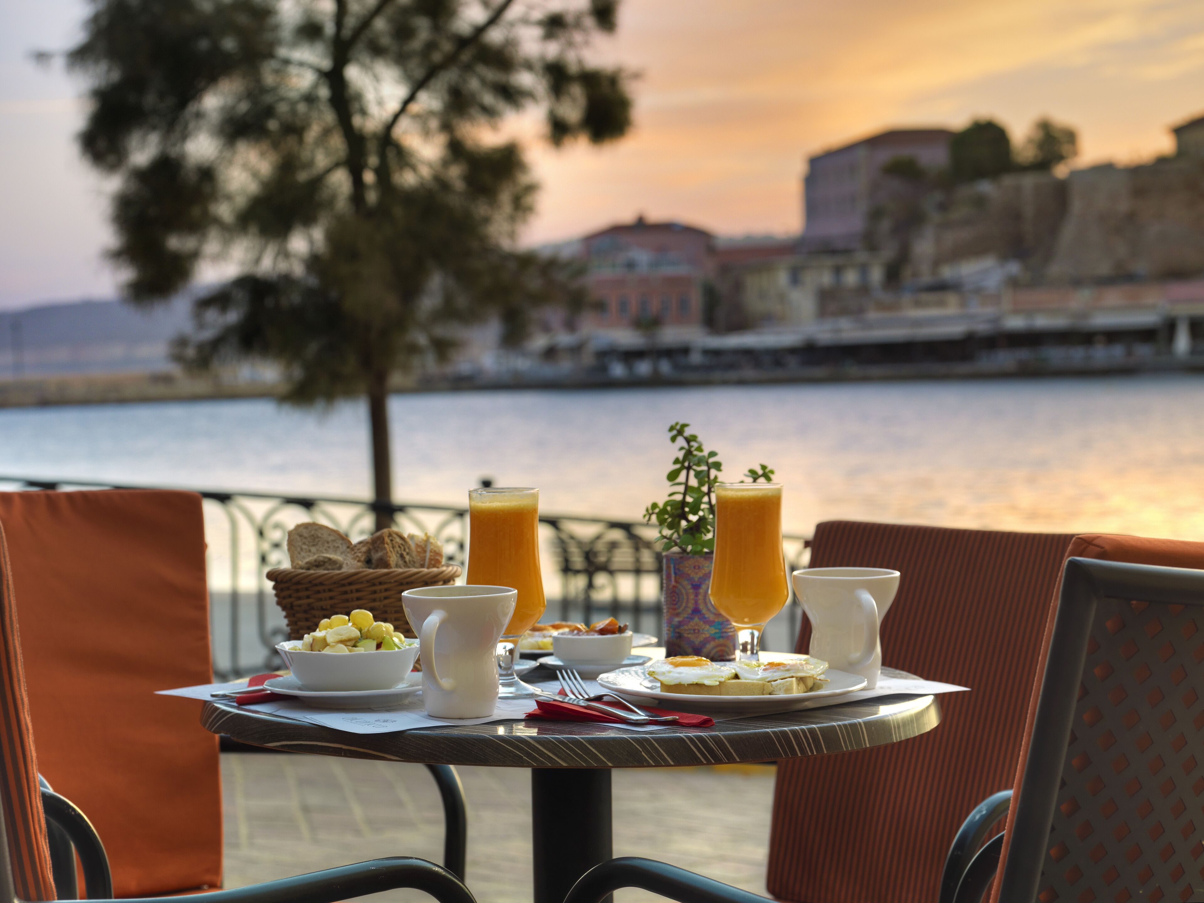 Una tavola imbandita con prodotti per la colazione, tra cui pane tostato, frutta e bevande, con vista su un'acqua e edifici sullo sfondo.