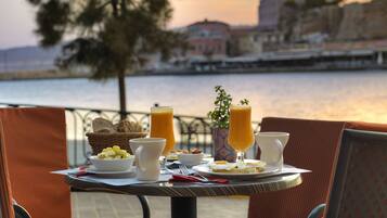 A table with breakfast items, including toast, fruit, and drinks, overlooking a body of water with buildings in the background.