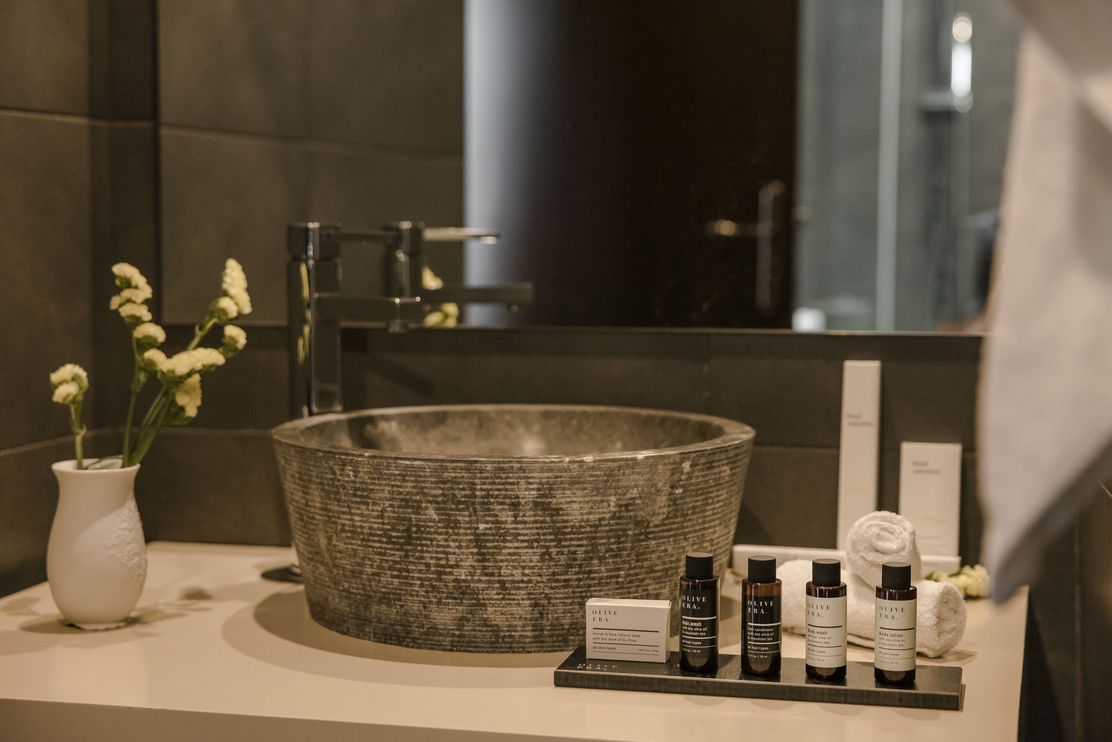 A bathroom vanity with a large, textured basin, a white vase with flowers, and a display of olive oil bottles.