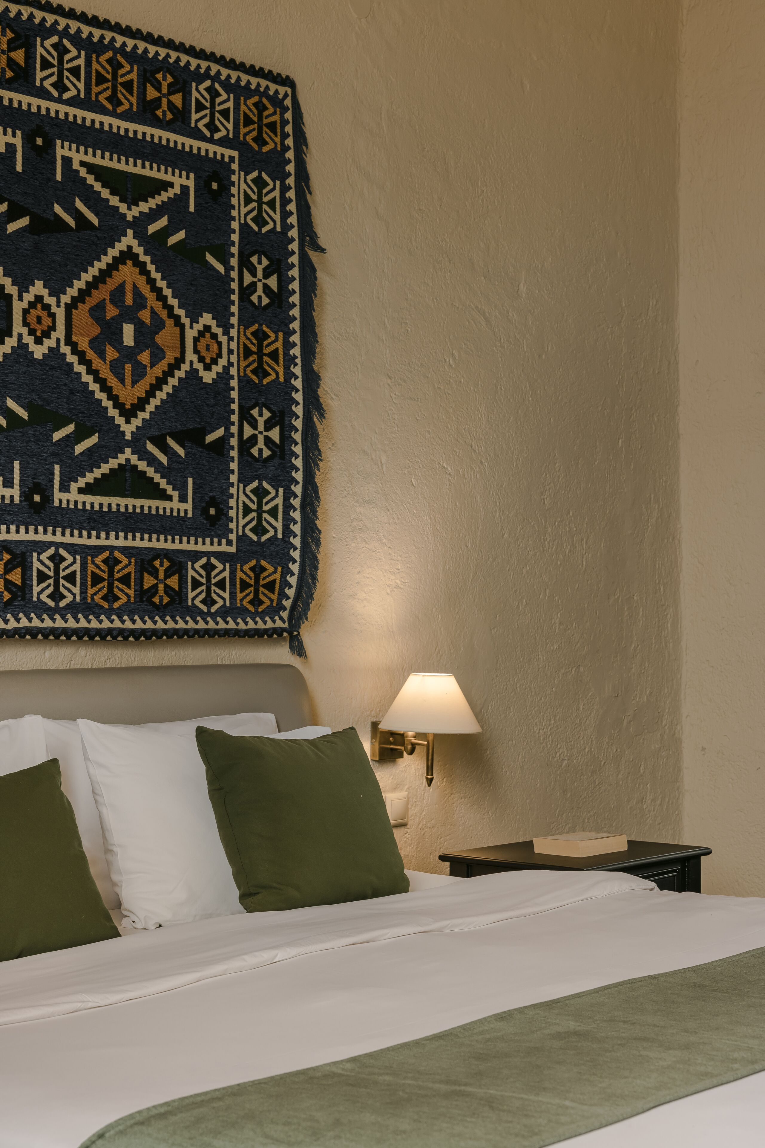 Una cama bien hecha con almohadas blancas y verdes, una manta estampada y una lámpara fijada a la pared.