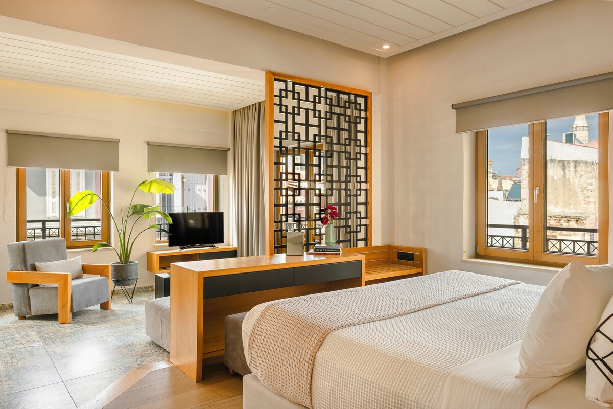 A modern hotel room with a bed, a desk, a chair, a TV, and a view of the city.