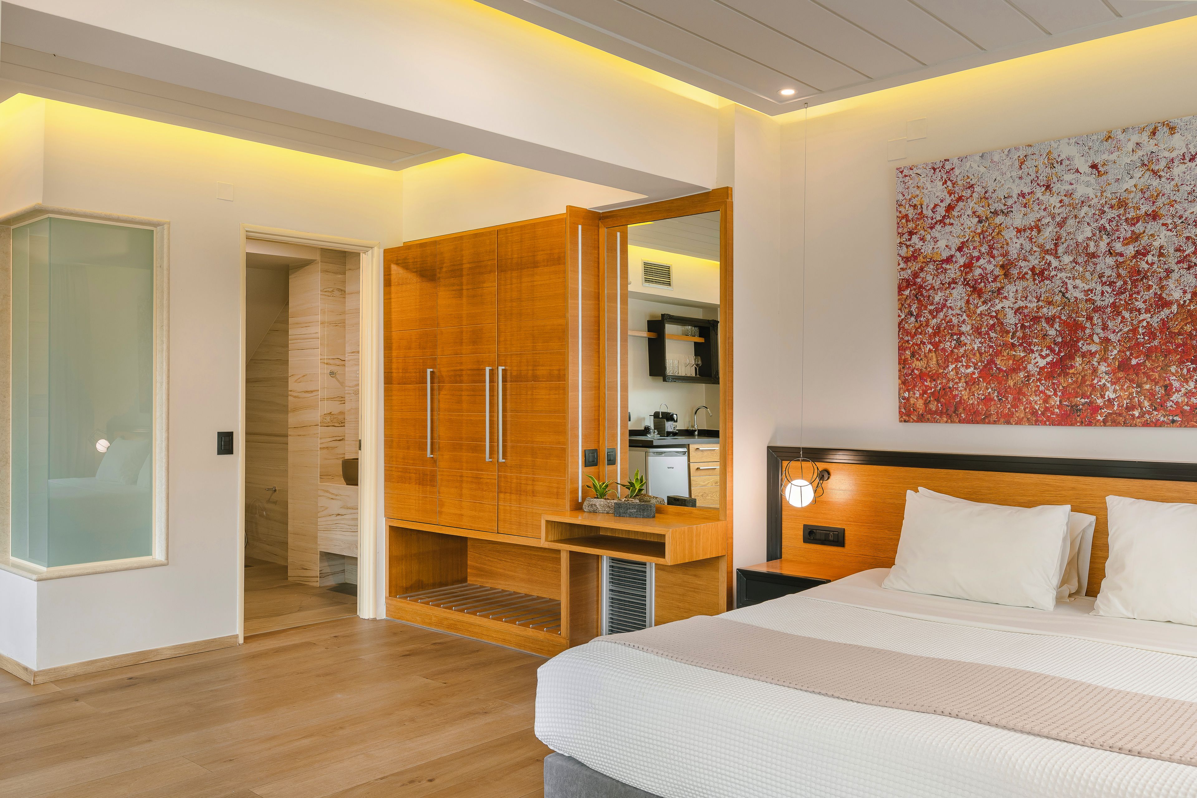 A modern bedroom with a large bed, a wooden wardrobe, a small kitchenette, and a bathroom with a glass door.