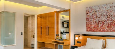 A modern bedroom with a large bed, a wooden wardrobe, a small kitchenette, and a bathroom with a glass door.