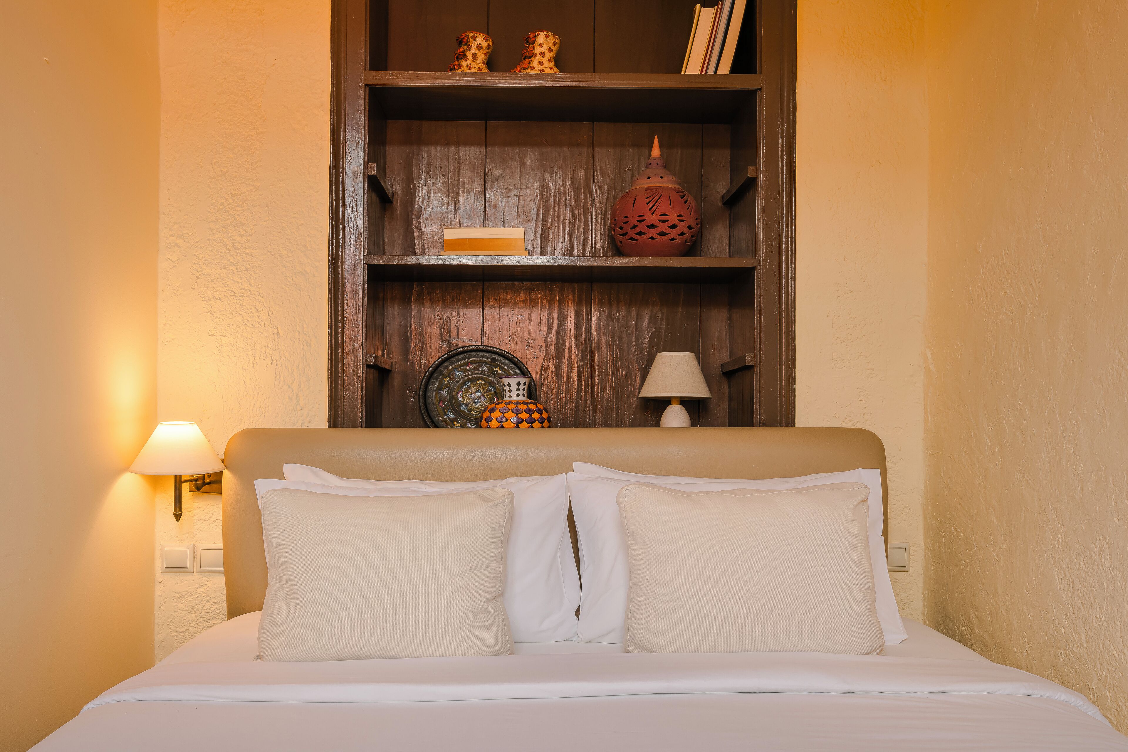 A neatly made bed with white linens and two pillows, a wooden shelf above with decorative items, and a wall-mounted lamp to the left.