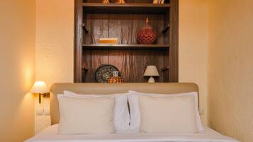 A neatly made bed with white linens and two pillows, a wooden shelf above with decorative items, and a wall-mounted lamp to the left.
