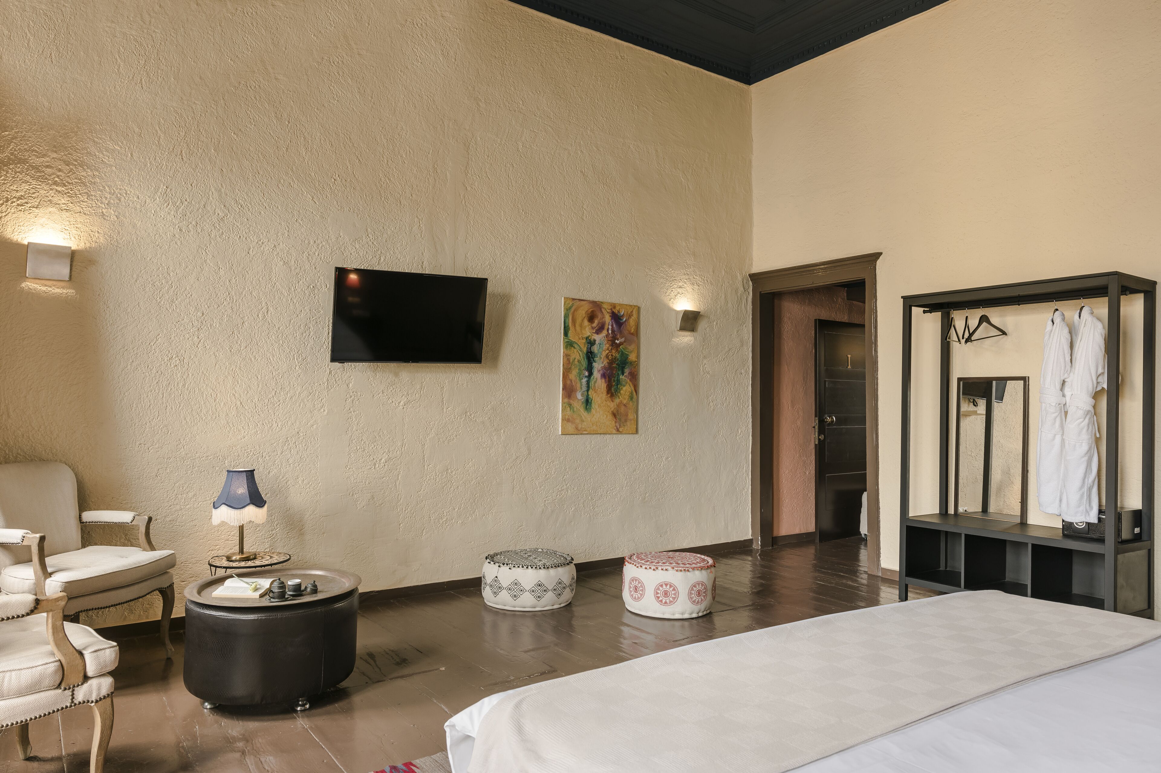A hotel room with a bed, a chair, a table with a lamp, a TV mounted on the wall, and a wardrobe with clothes hanging inside.