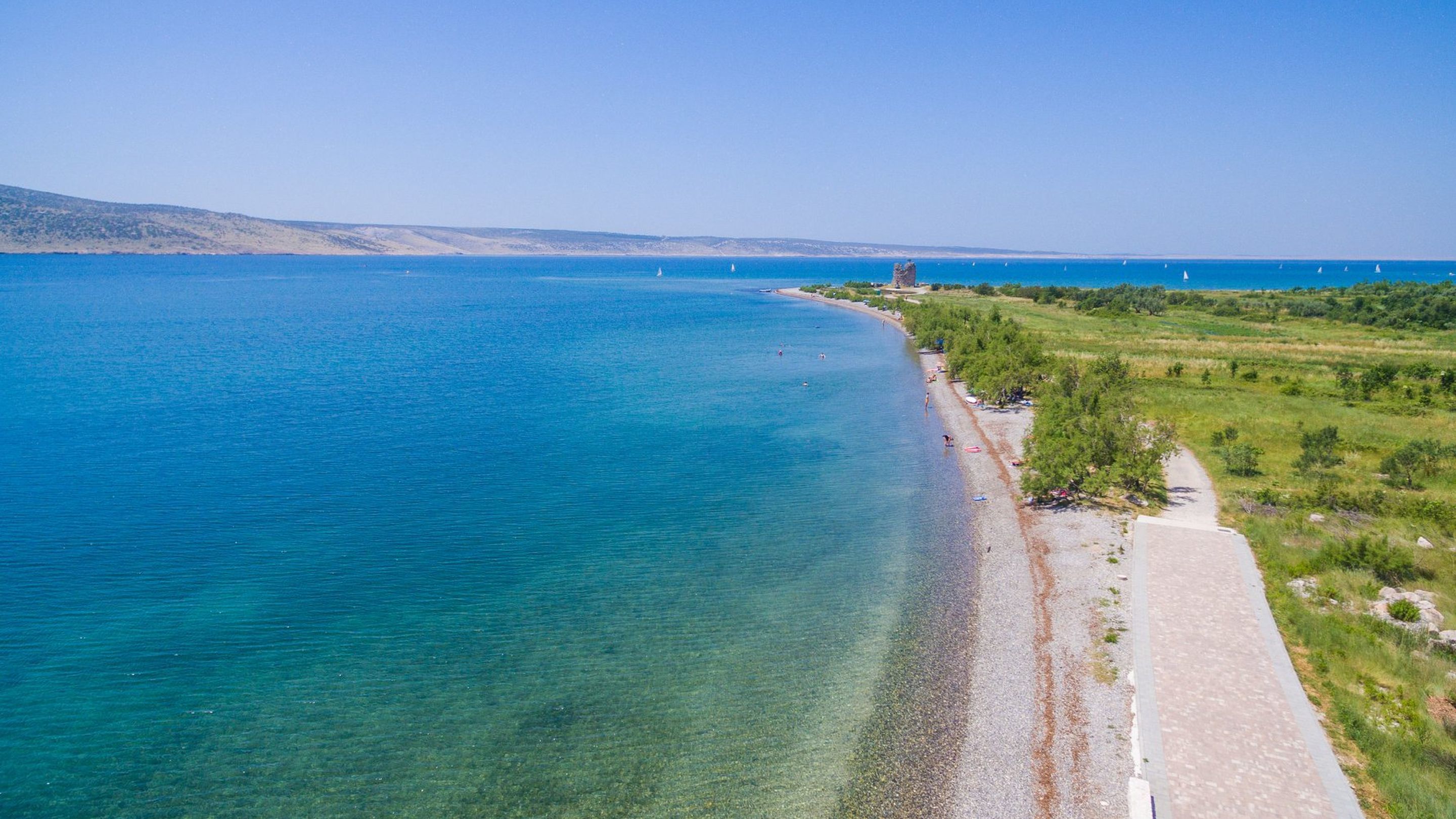 Uma estrada costeira com uma praia à direita, águas azuis cristalinas e colinas ao longe.