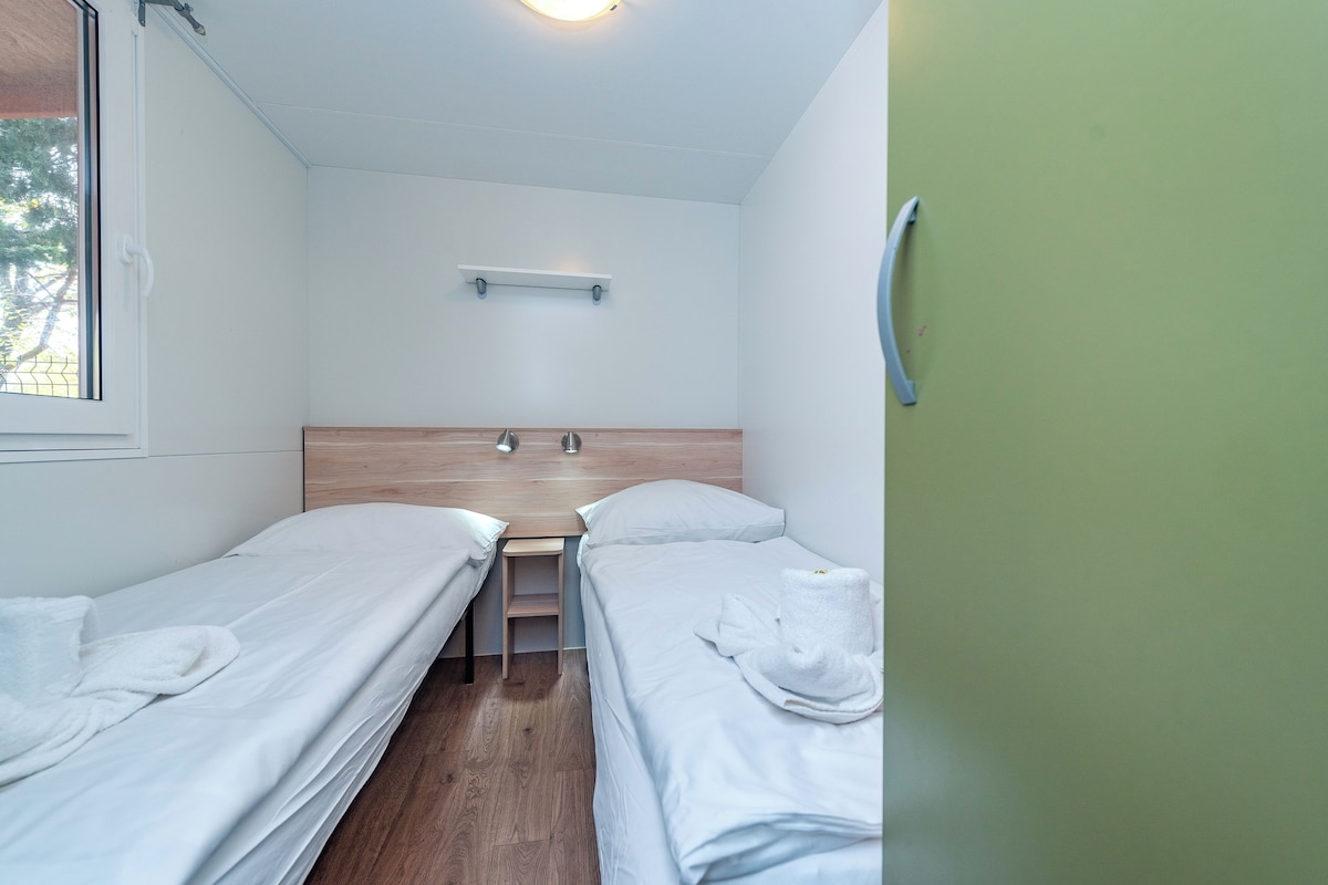 A small, clean room with two single beds, a wooden desk, and a small shelf above the desk.
