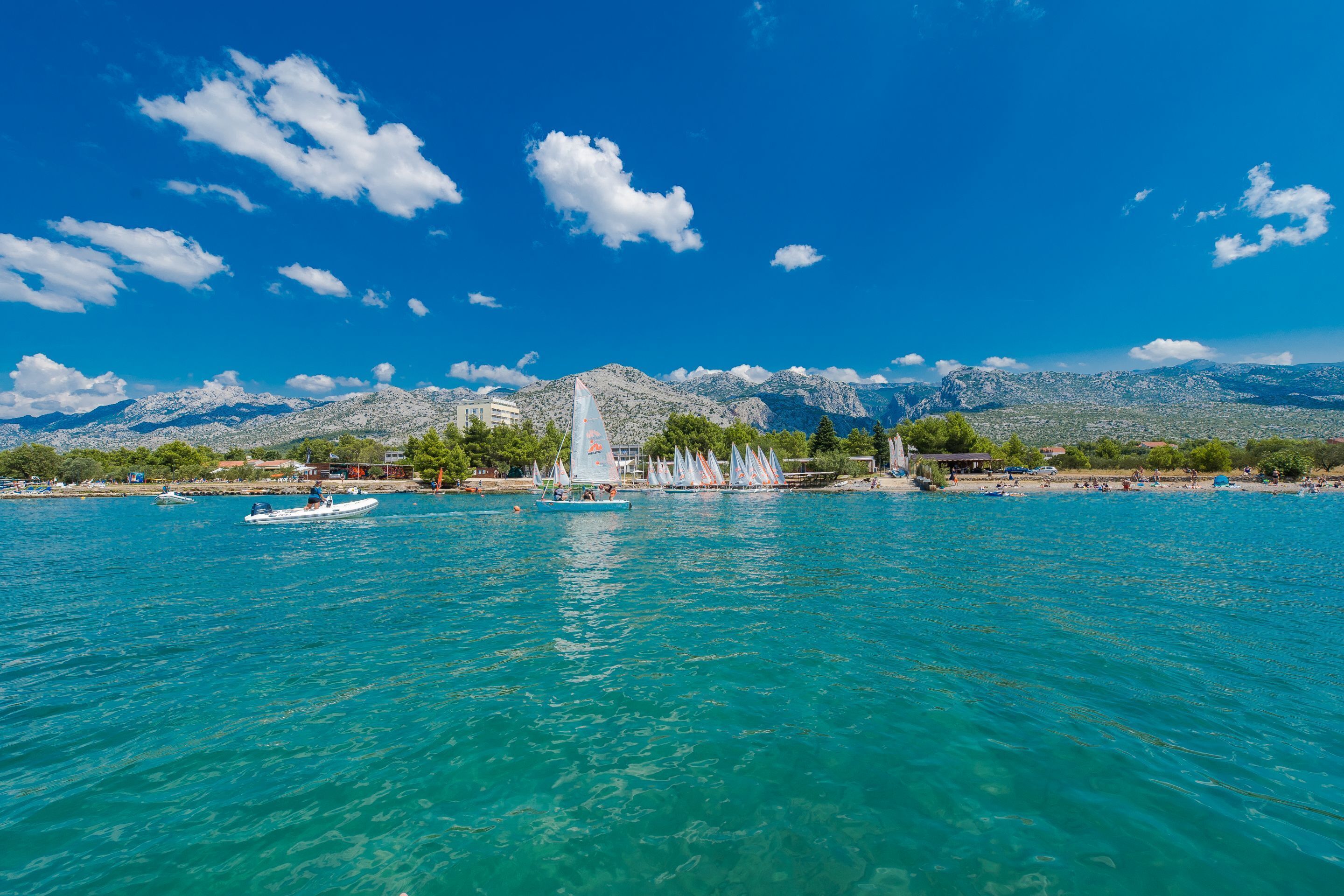 A coastal scene with a clear blue sky, a sailboat, and a mountainous backdrop.