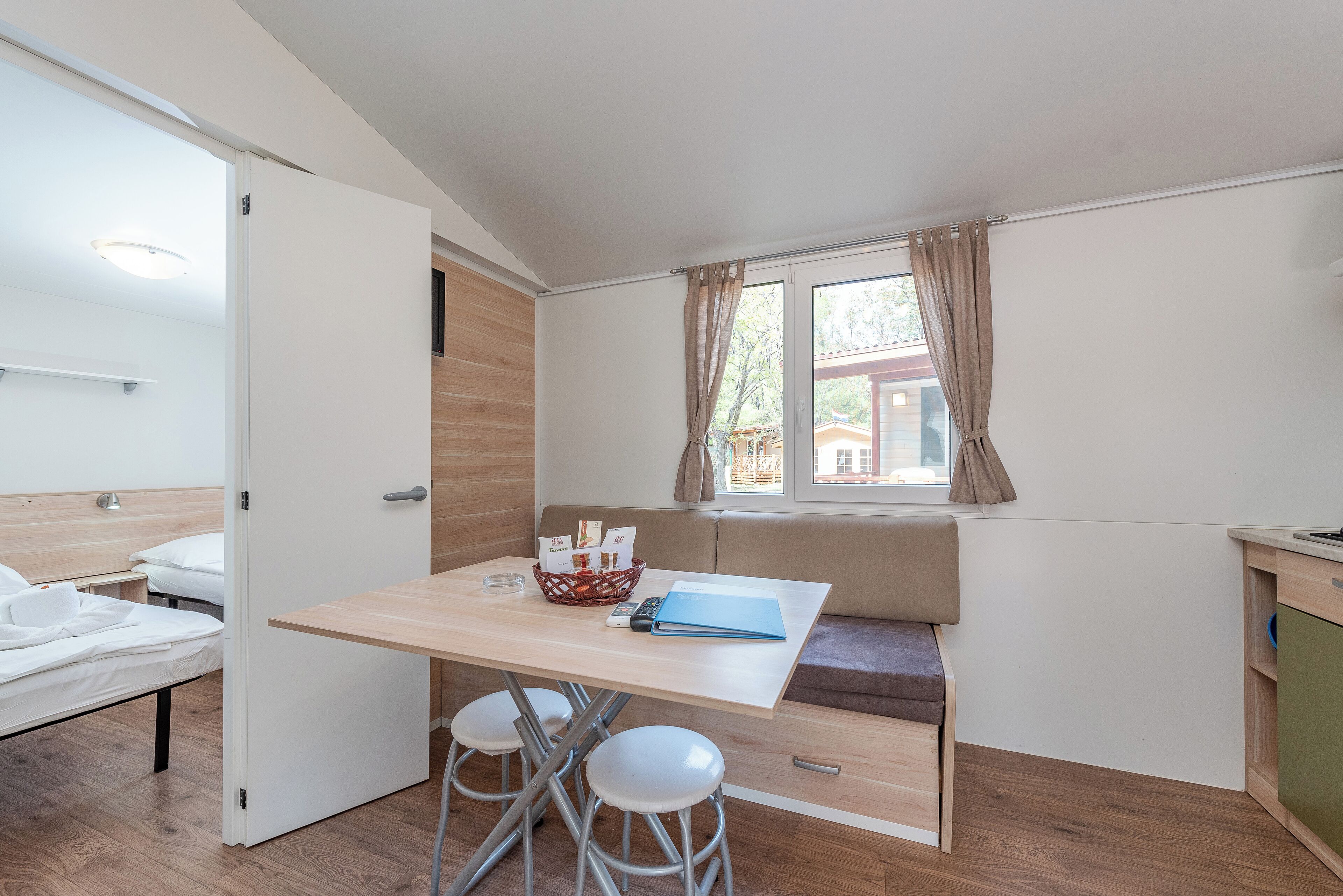 A compact room with a bed, a dining table with stools, a bench, and a window with curtains.