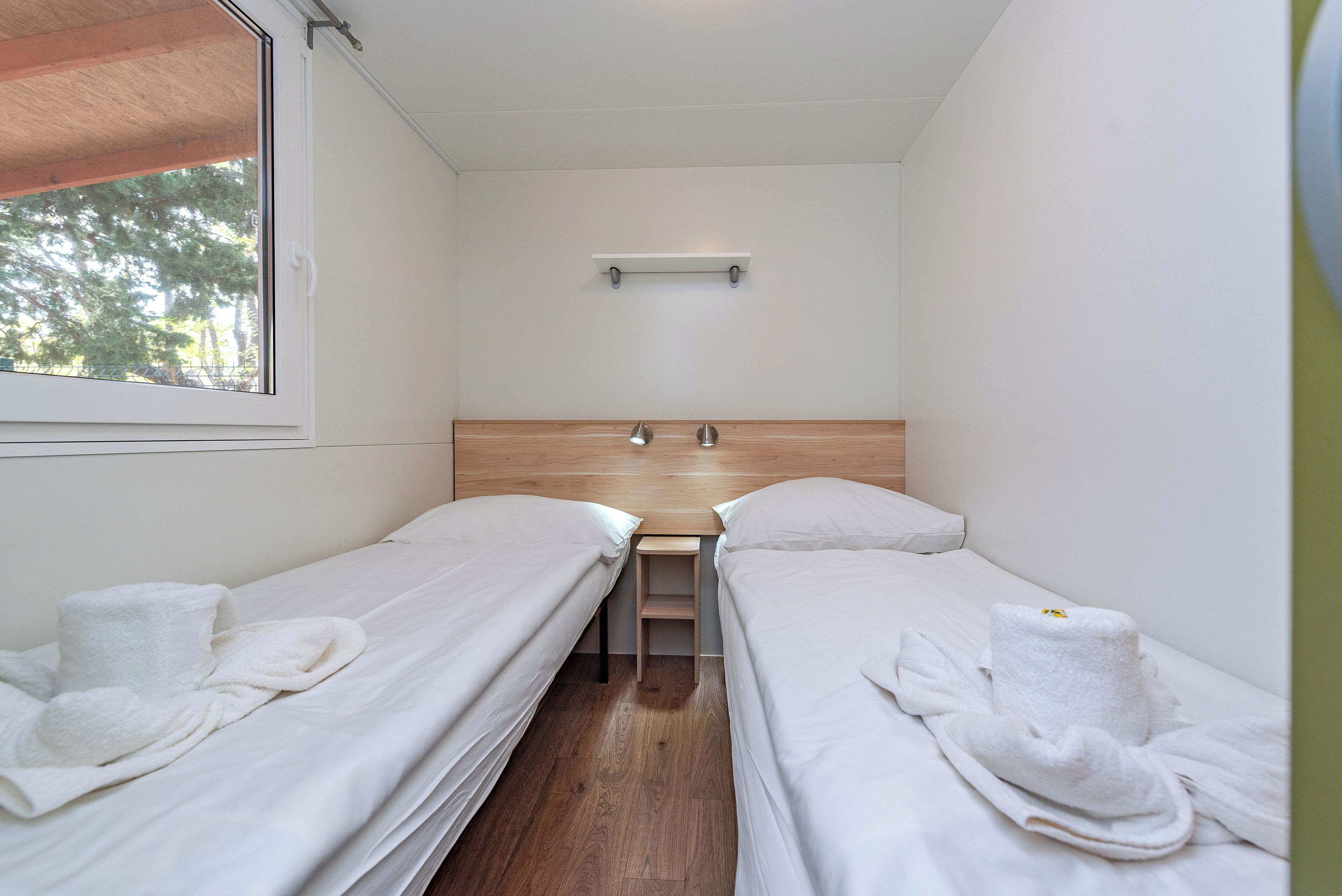 A small, clean room with two single beds, a small table, and a shelf above the table.