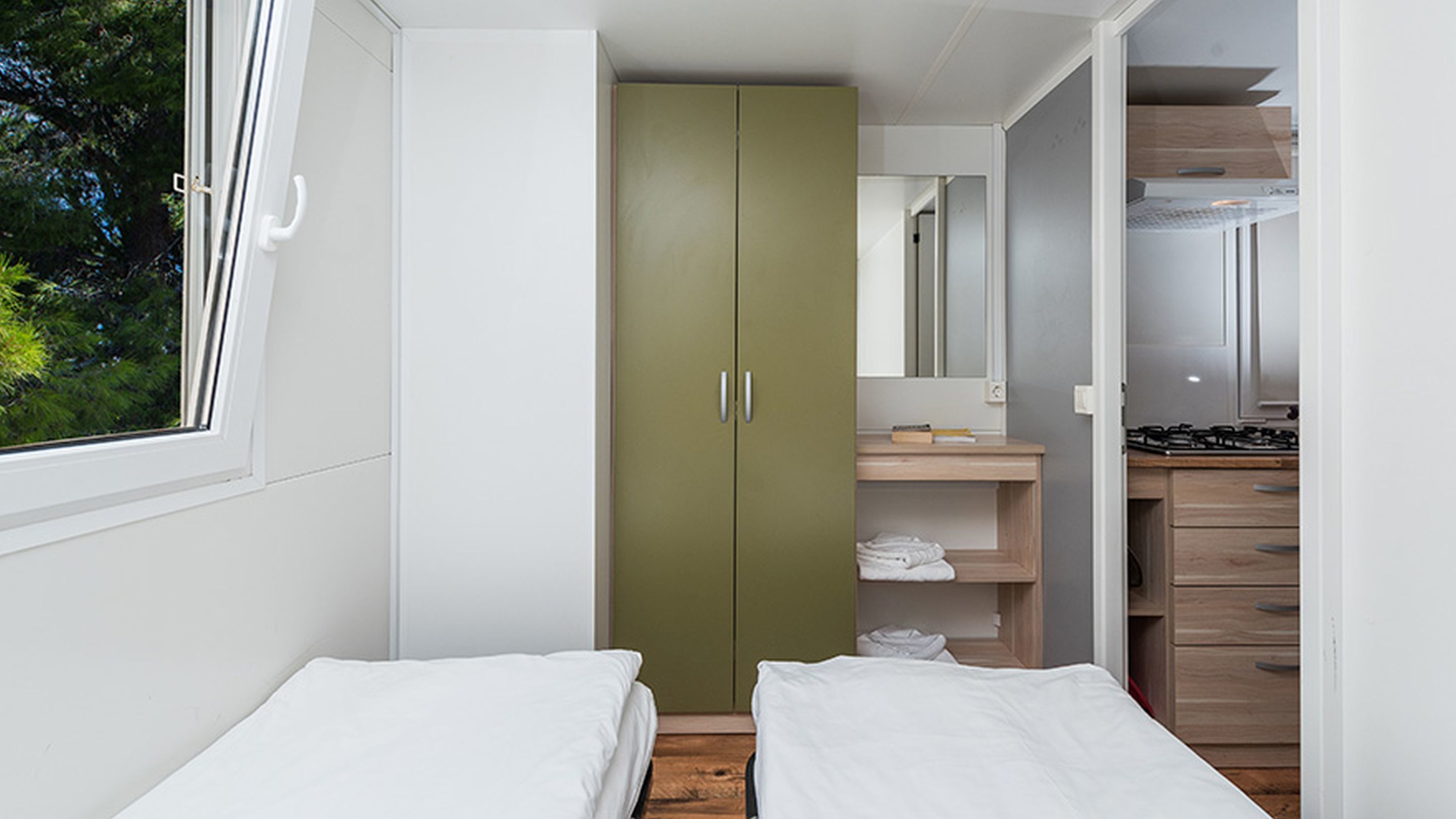 A bedroom with two beds, a green wardrobe, a small bathroom with a mirror, and a kitchenette.