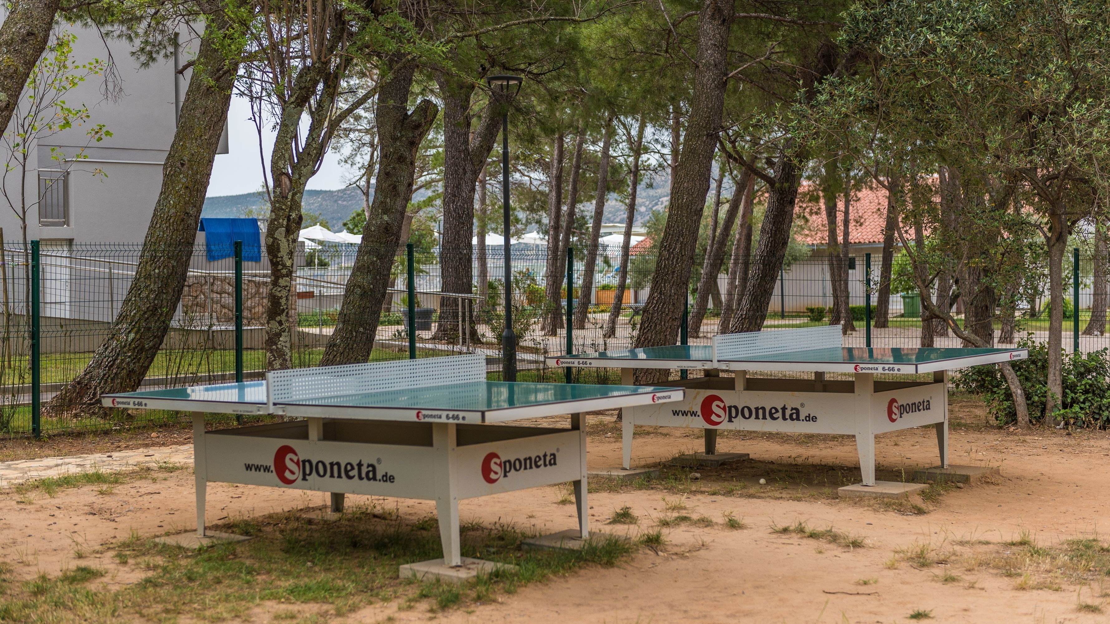 Deux tables de ping-pong de la marque « Sponeta », entourées d’arbres et d’une clôture.