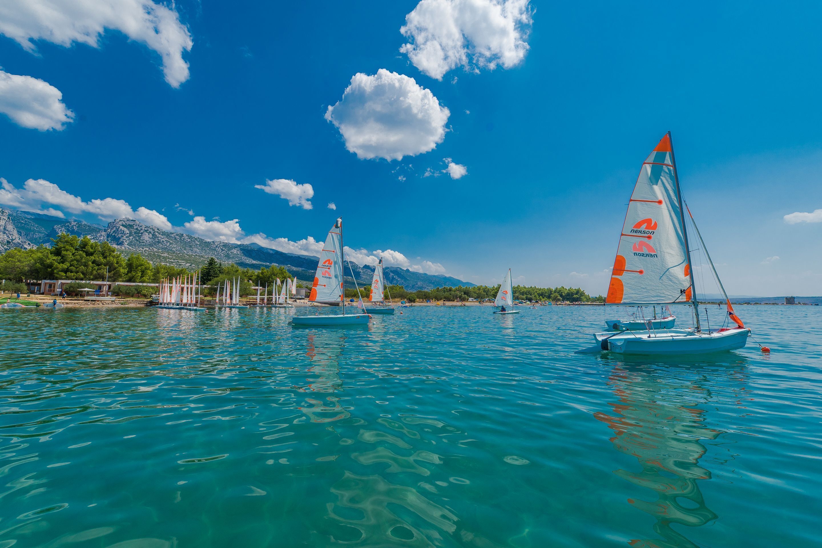 A calm lake with sailboats, clear blue sky, and mountains in the background.