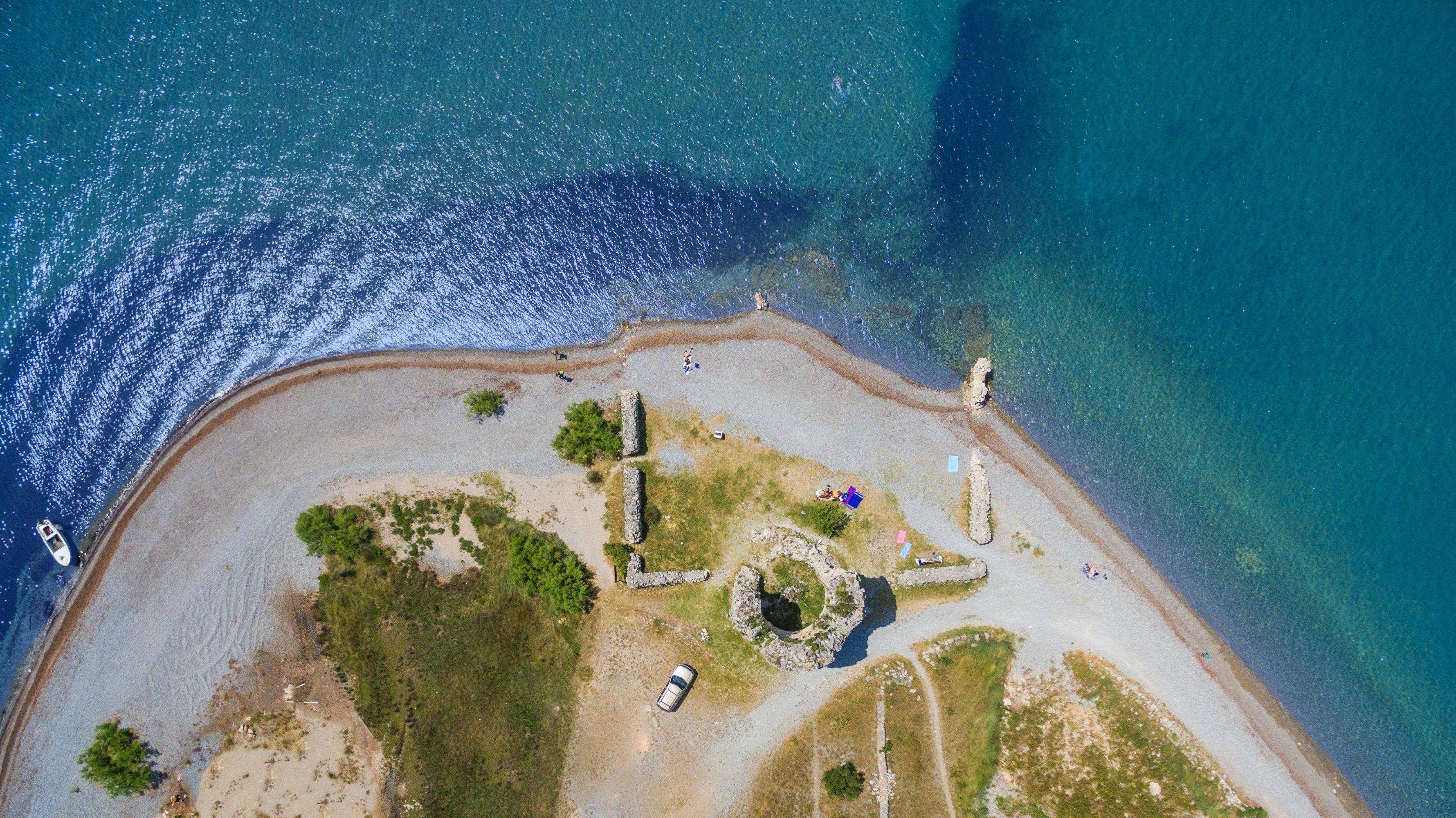 An aerial view of a coastal area with a sandy beach, a small rocky outcrop, and a few scattered trees.