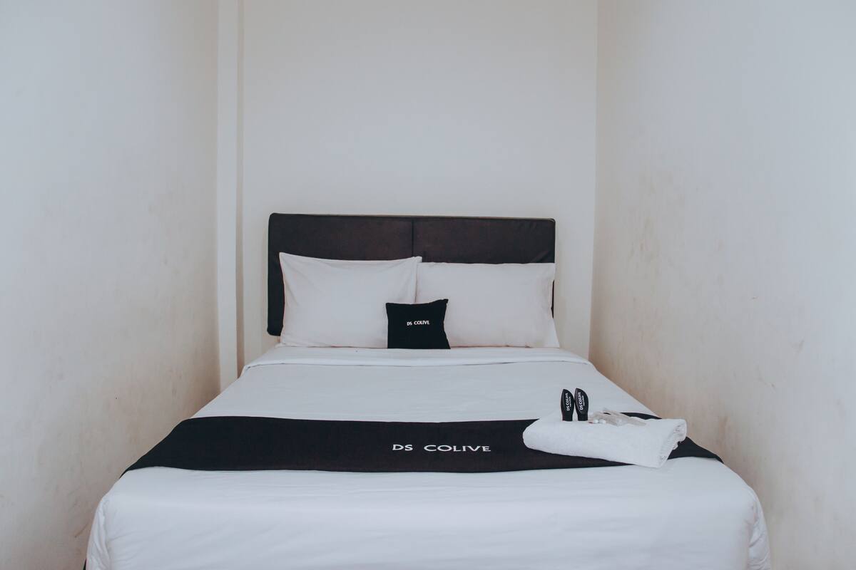 Una cama bien hecha con un cabecero negro y ropa de cama blanca, una almohada blanco y negro, y una toalla de la marca DS COLIVE.