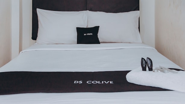 A neatly made bed with a black and white bedding set, a black pillow with "DS COLIVE" text, and a pair of black shoes on the bed. - DS Residence Siliwangi (Semarang)